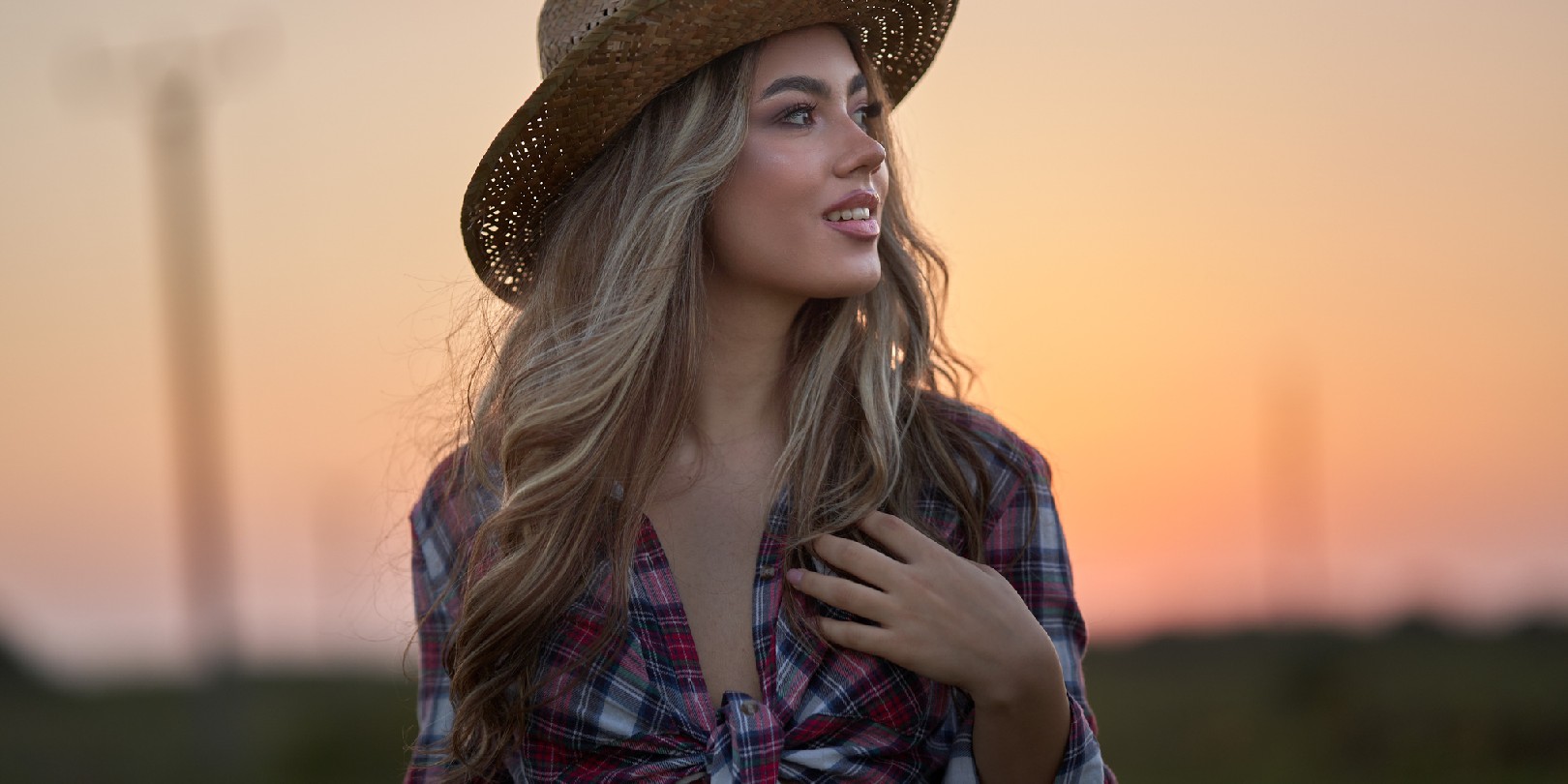 Beautiful cowgirl in hat, plaid shirt and short jeans at sunset in a countryside field