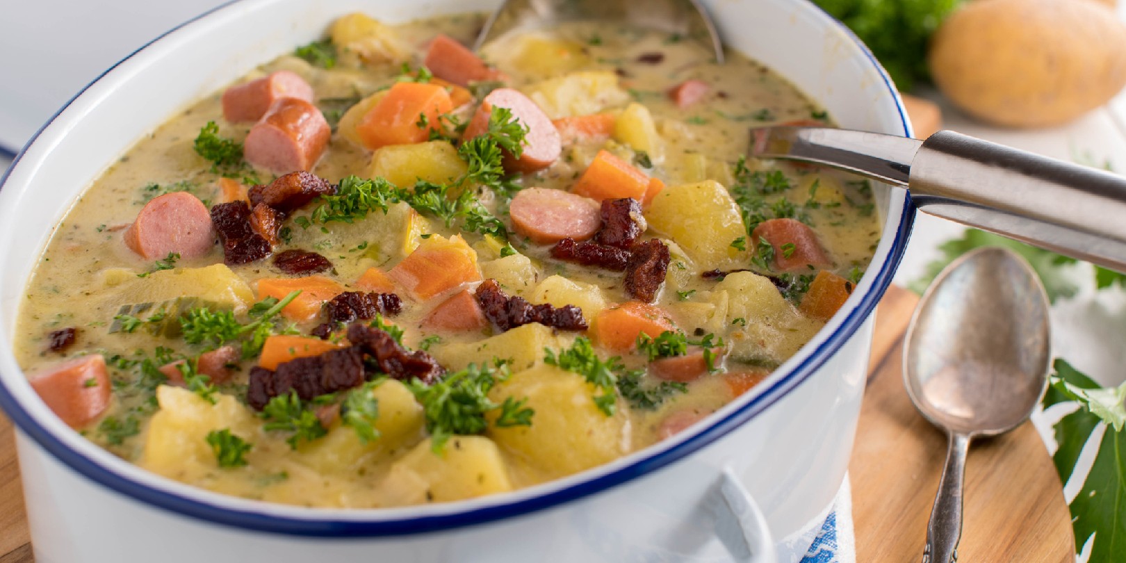 Homemade potato soup or potato stew with crispy bacon, vienna sausage and vegetables. Served in old fashione enamel pot on kitchen table background